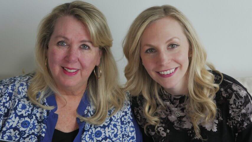 Two women smiling, wearing floral tops, sitting together indoors; blonde hair, friendly expression, casual setting.