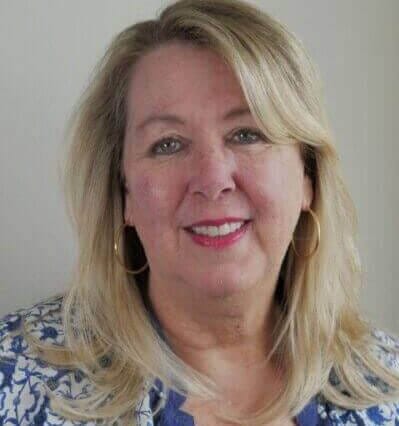 Senior woman with blonde hair and blue floral blouse smiling at camera, wearing gold hoop earrings, professional headshot.
